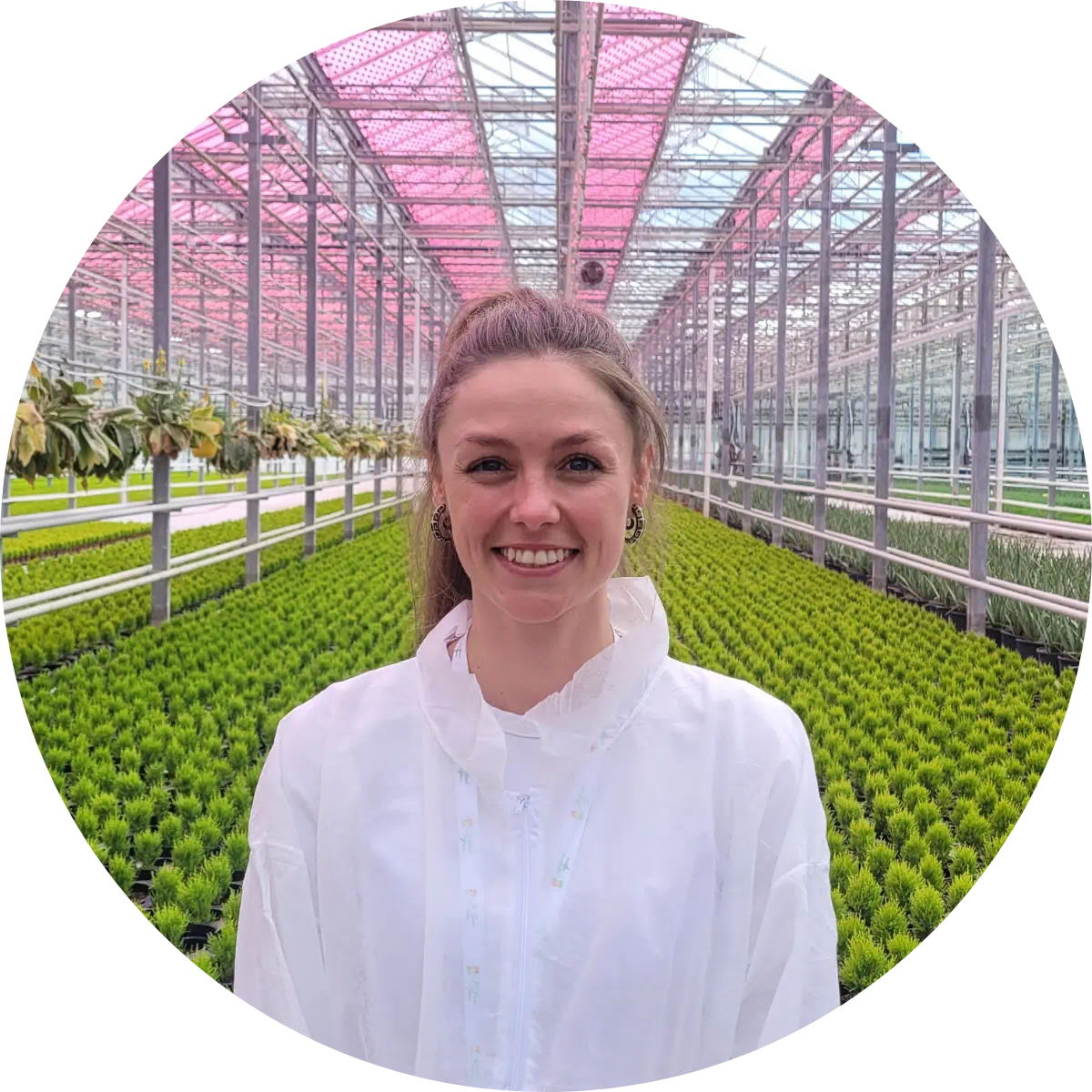 Portrait of Dr. Delaney Nash standing in a greenhouse with rows of young plants, wearing protective lab clothing in a controlled growing environment.